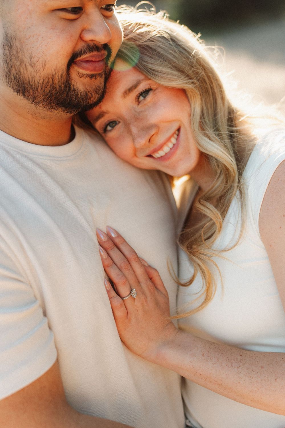 Smiling blonde woman leans against bearded man, showing engagement ring, in warm golden light.