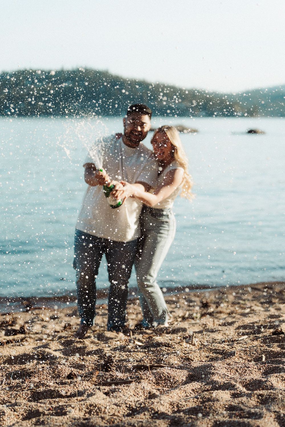 Couple celebrating on a sandy beach by popping champagne bottle with sparkling wine spraying into the air.