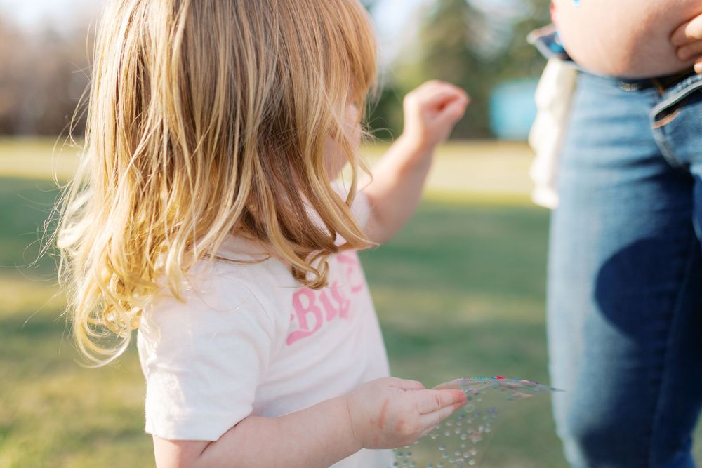 Toddler girl with blonde hair playing outdoors in a park, wearing a white shirt on a sunny day.