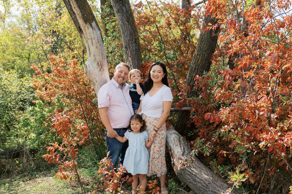 Family of four posing by a large tree surrounded by vibrant red and green autumn foliage outdoors.