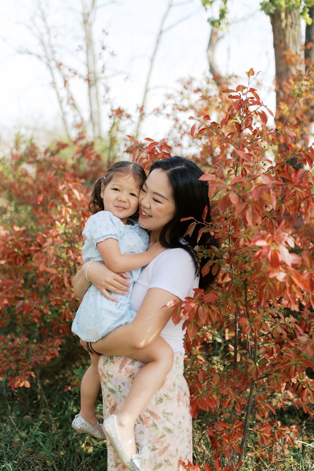 A mother holds her young daughter among vibrant red autumn leaves, sharing a tender embrace outdoors.
