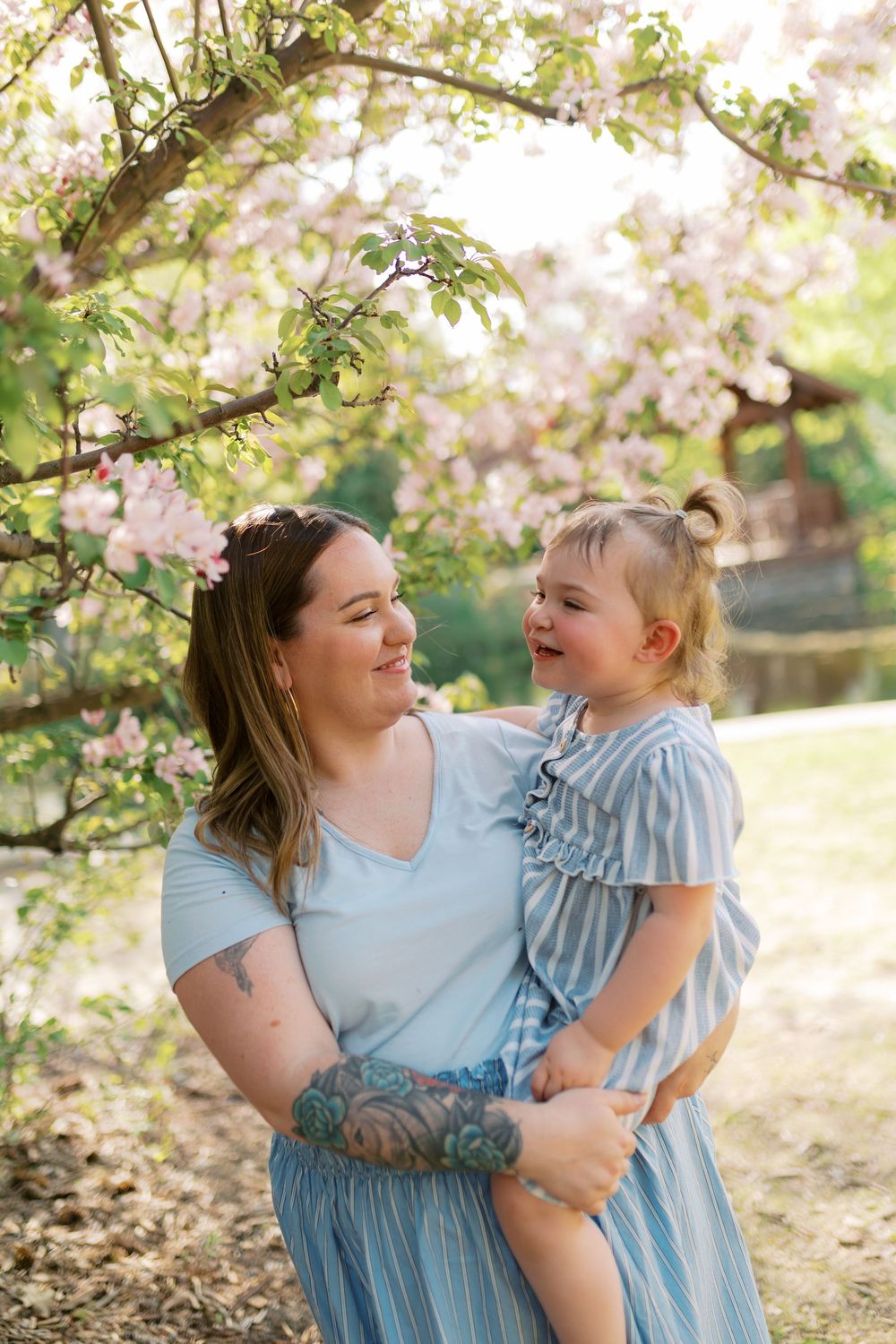 A tattooed mother holds her smiling toddler daughter under blooming pink cherry blossom trees, both wearing light blue outfits.