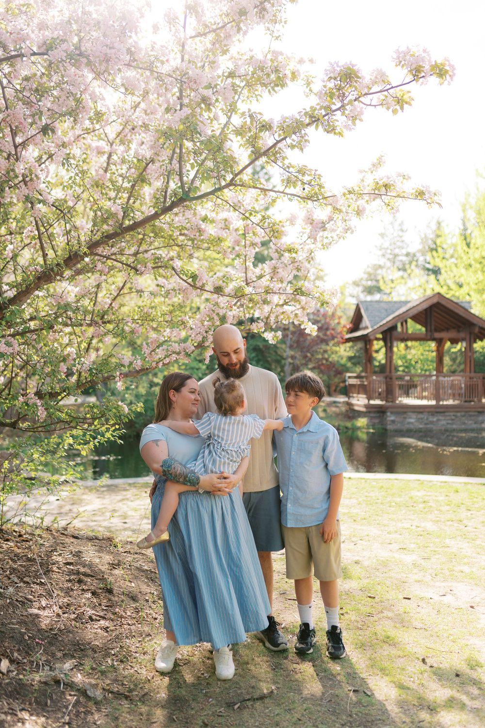Family of four enjoying a sunny spring day under blooming pink trees in a peaceful garden park.