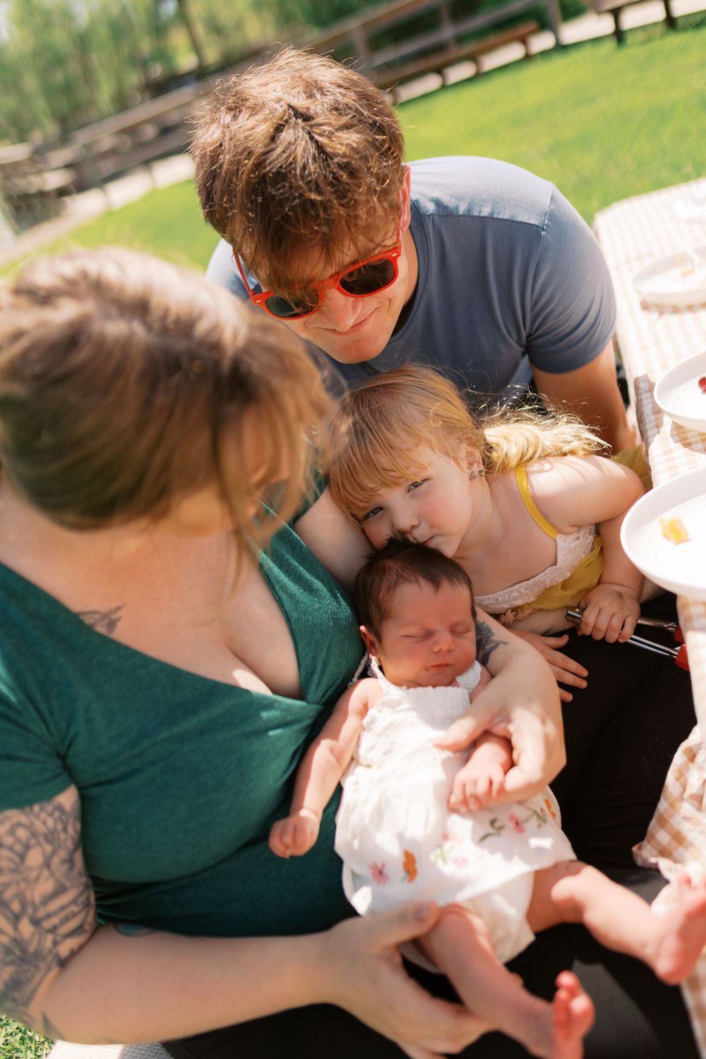 Family gathered outdoors admiring a newborn baby, with a toddler girl, tattooed mother, and father wearing sunglasses.
