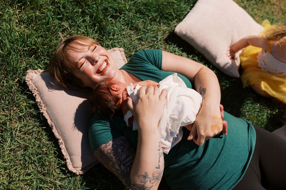 A smiling woman with tattoos lies on grass with a pillow, playfully holding a baby on her chest outdoors.