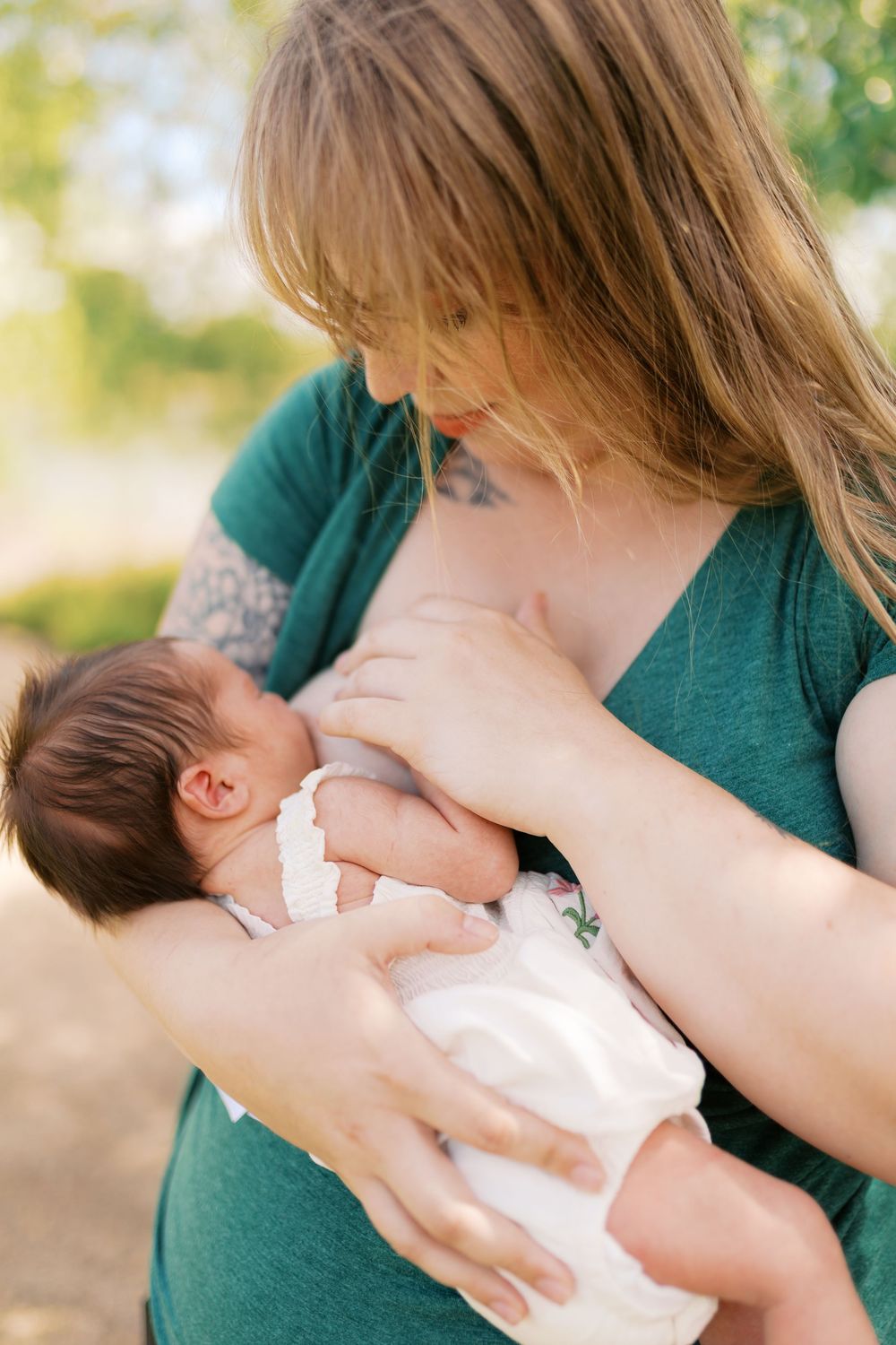 A mother in a green shirt tenderly breastfeeds her newborn baby outdoors in natural light.