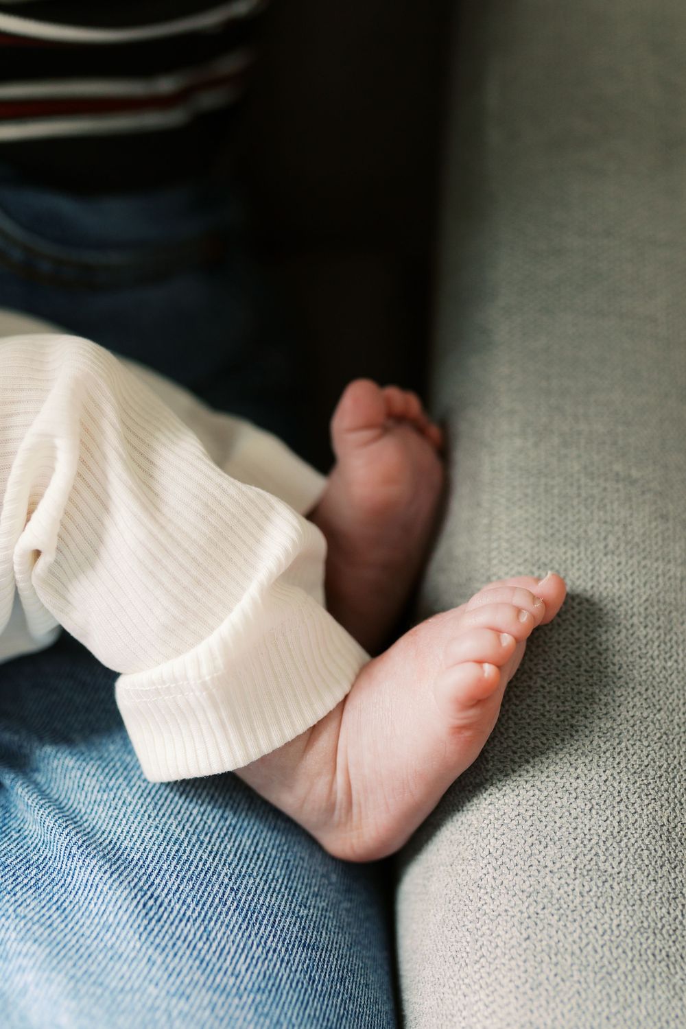 Close-up of tiny newborn baby feet resting on a parent's lap, wearing white ribbed pants on a gray sofa.