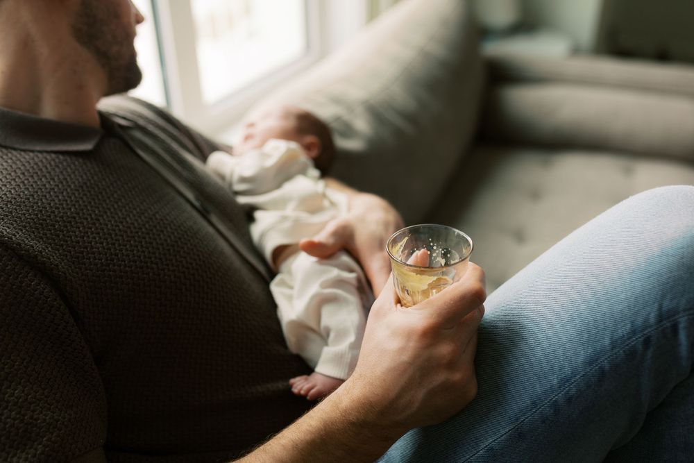 Father holding a glass of whiskey while cradling a sleeping newborn baby on a couch at home.