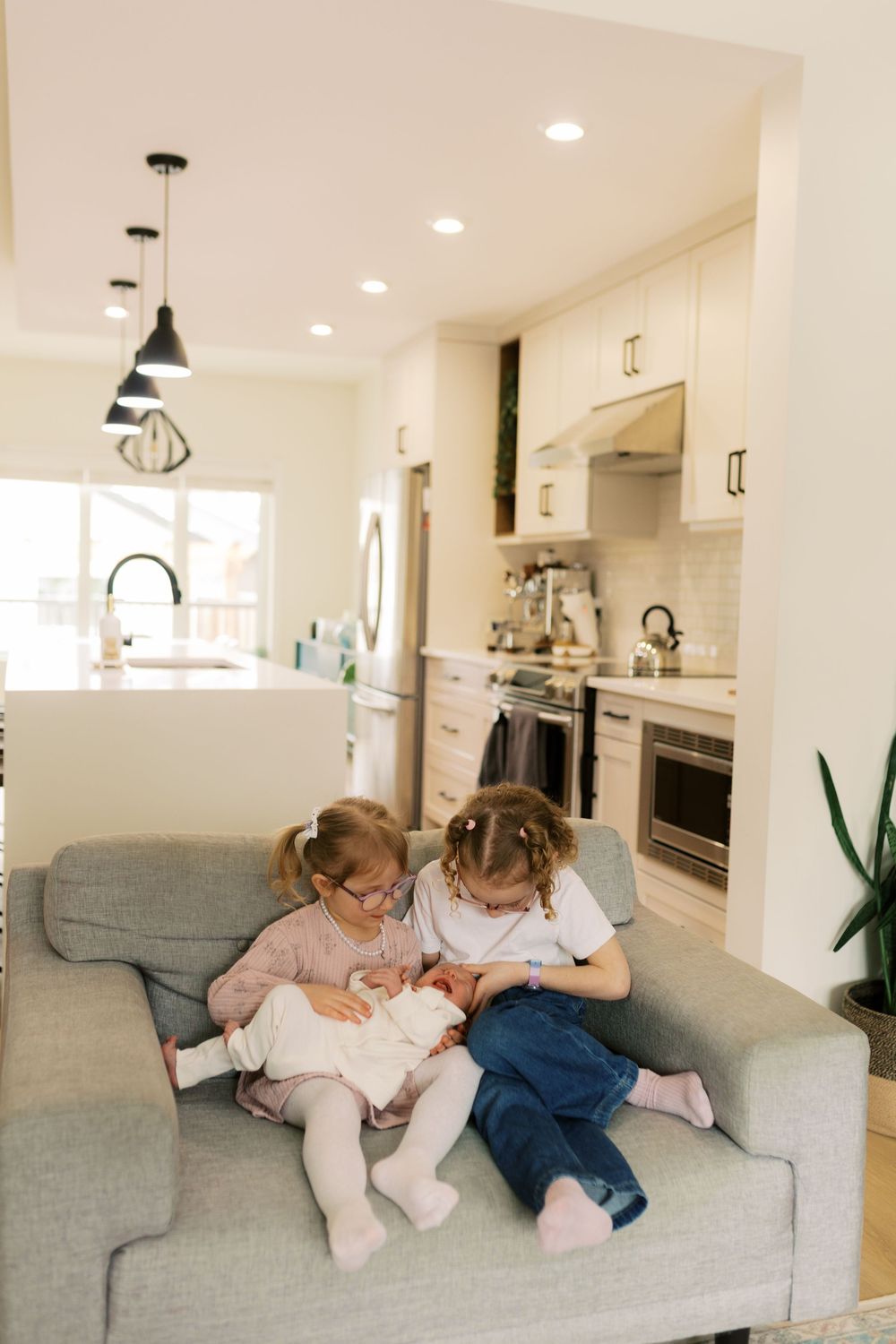 Two young girls sit together on a gray sofa in a bright modern kitchen, looking at a baby doll.