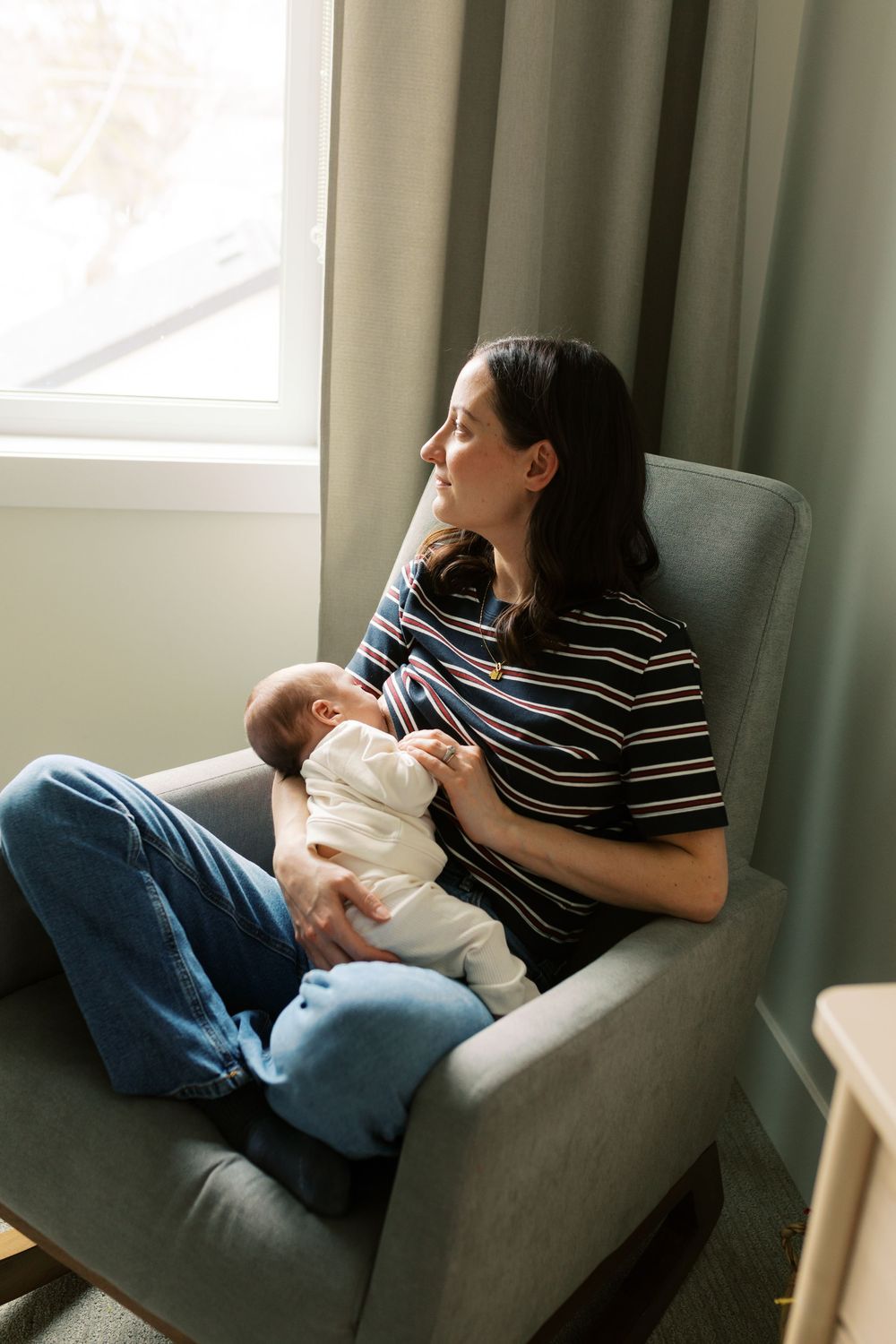 Mother in striped shirt breastfeeding newborn baby while sitting in gray rocking chair by window.