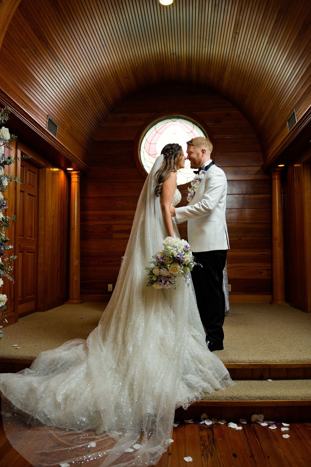 Bride and groom share a romantic moment in a wooden chapel with arched ceiling, stained glass window, and long bridal veil.
