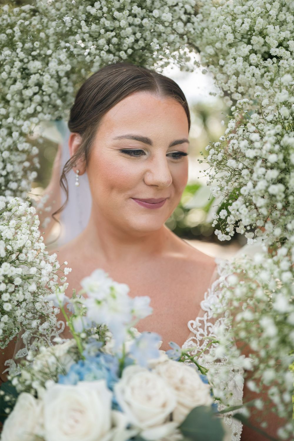 Bride surrounded by baby's breath flowers holding a bouquet of white roses and blue hydrangeas on her wedding day.