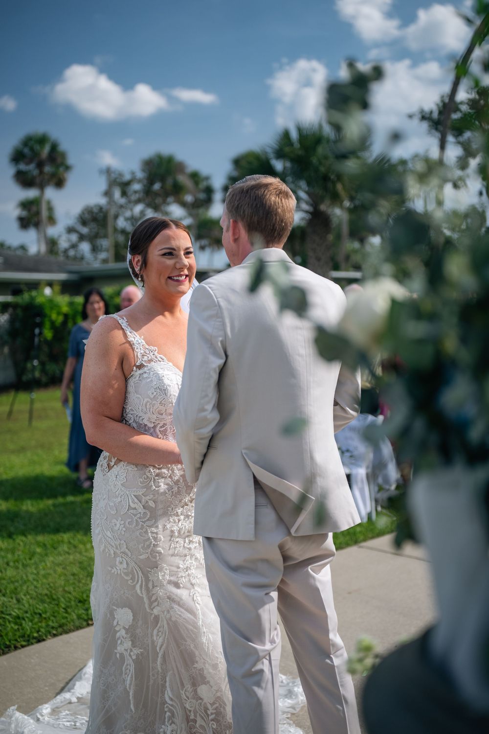 Bride in lace gown smiles at groom in tan suit during outdoor wedding ceremony with palm trees.