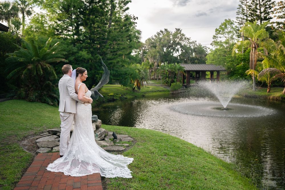 Bride and groom stand by a serene pond with a fountain, surrounded by lush gardens at an outdoor wedding venue.