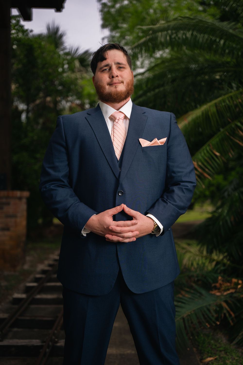 Man in navy blue suit with pink tie and pocket square posing outdoors near tropical palm trees.