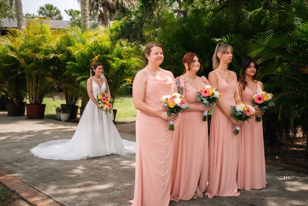 Bride in white gown stands apart from three bridesmaids in blush pink dresses holding colorful bouquets outdoors.