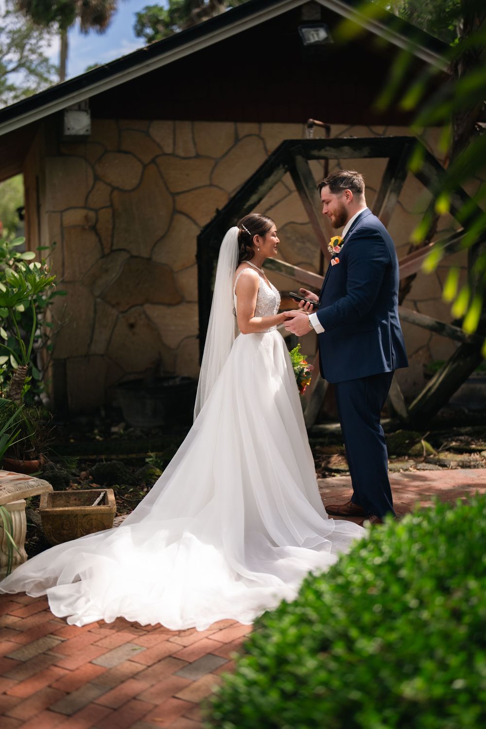 Bride and groom exchange vows outdoors beside a stone building, her long white veil trailing on brick pavers.