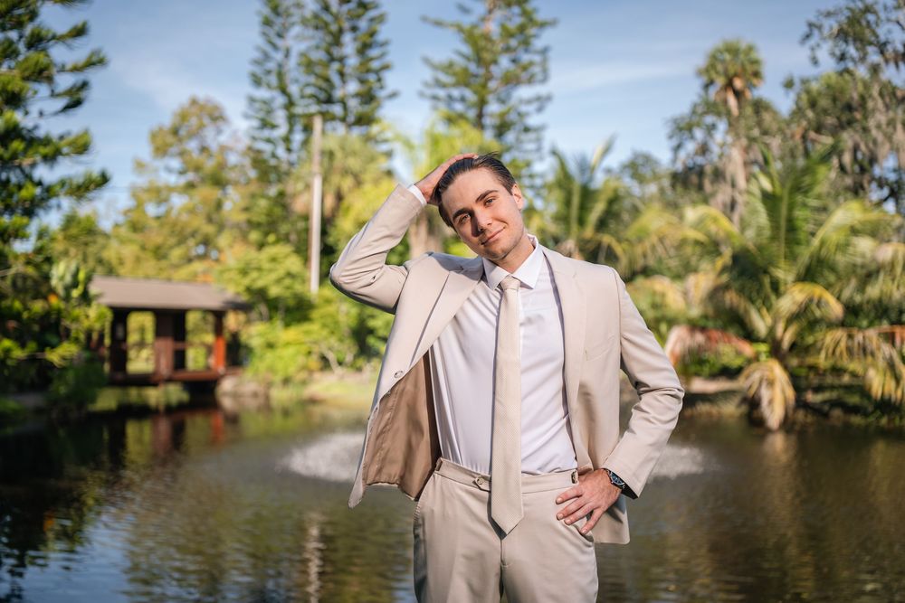 Man in beige suit posing confidently in a lush garden with a pond and fountain backdrop on a sunny day.