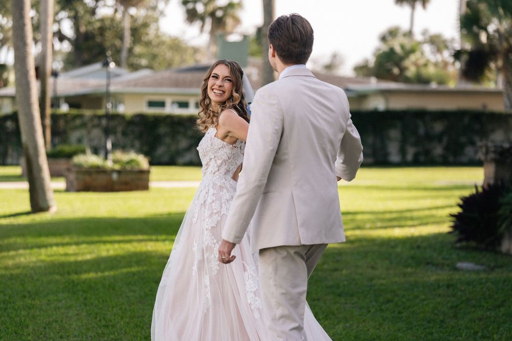 Bride in lace gown smiles at groom in tan suit while holding hands on green lawn at outdoor wedding.