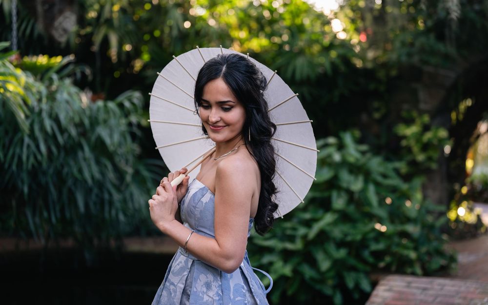 Young woman in elegant dress holding a white paper parasol umbrella, smiling in a lush green garden setting.