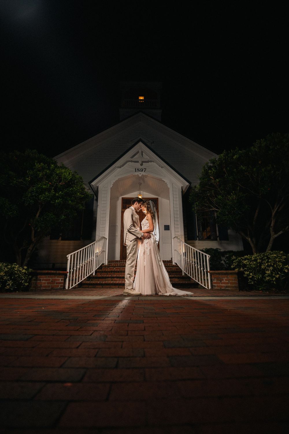 Bride and groom share a romantic kiss on church steps at night, illuminated by warm light in a stunning wedding portrait.