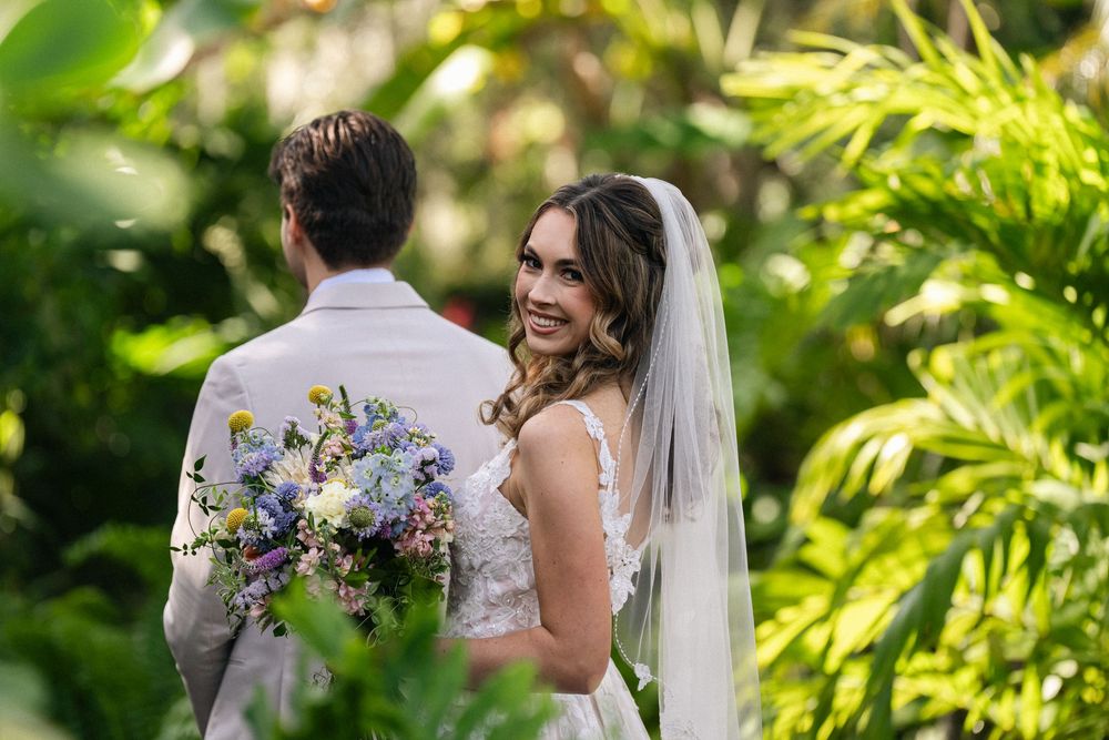Bride in white gown and veil smiles at groom holding colorful bouquet during outdoor wedding ceremony.