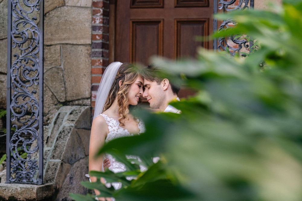 Bride and groom share a romantic moment framed by lush greenery near an ornate wooden church door.