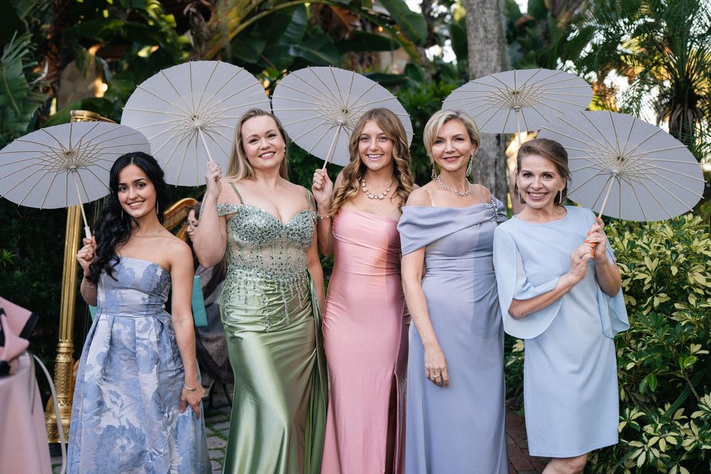 Five women in elegant pastel gowns pose with white parasols outdoors at a formal garden event.