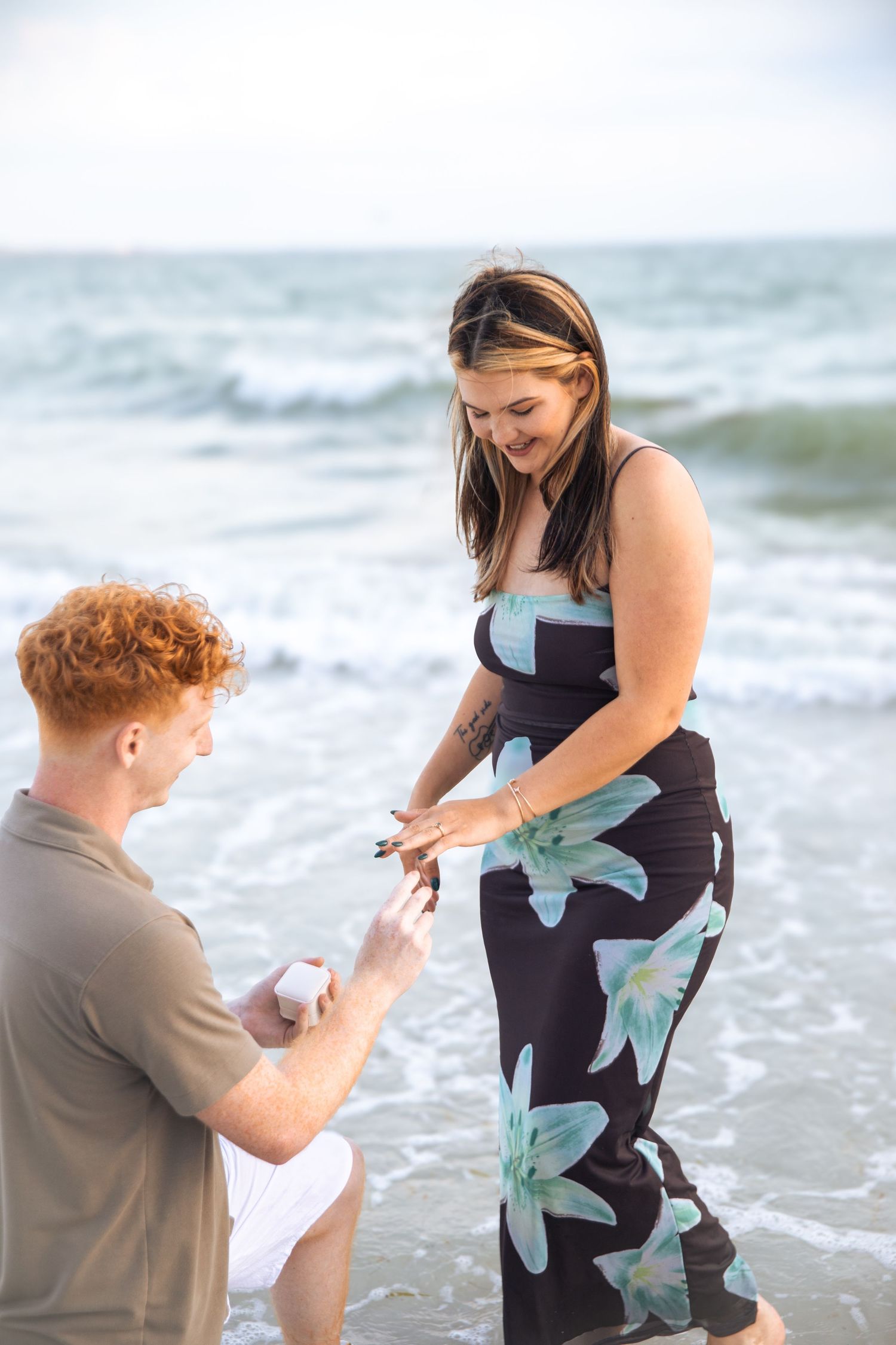 A red-haired man proposes on one knee to a smiling woman in a floral dress on a beach.