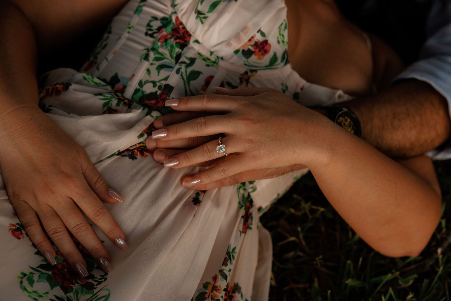 Close-up of couple holding hands showing engagement ring, woman wearing floral dress outdoors.