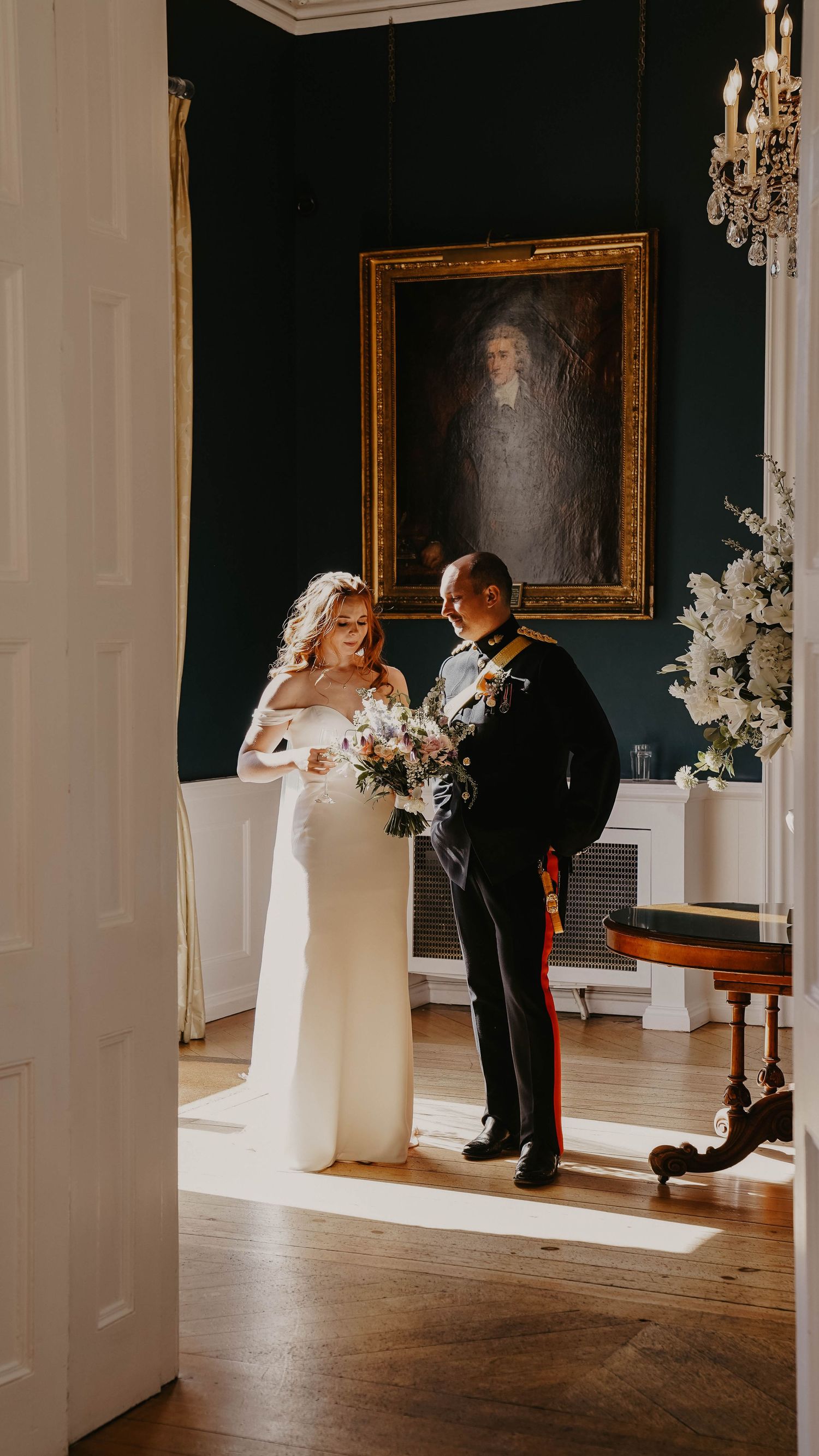 Bride in white gown and groom in military uniform exchange vows in elegant room with portrait and chandelier.