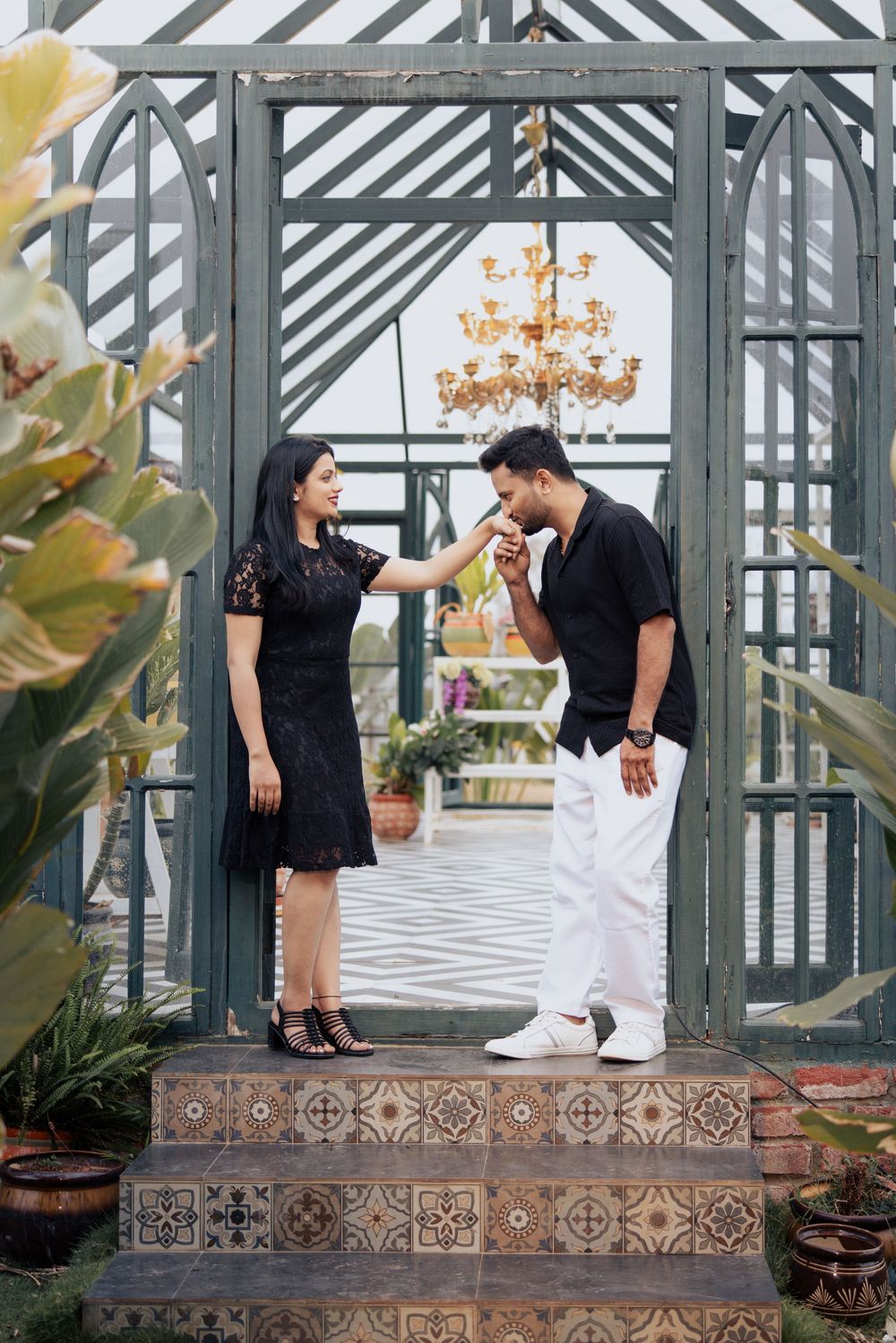 A man in black shirt and white pants kisses a woman's hand outside an elegant greenhouse.