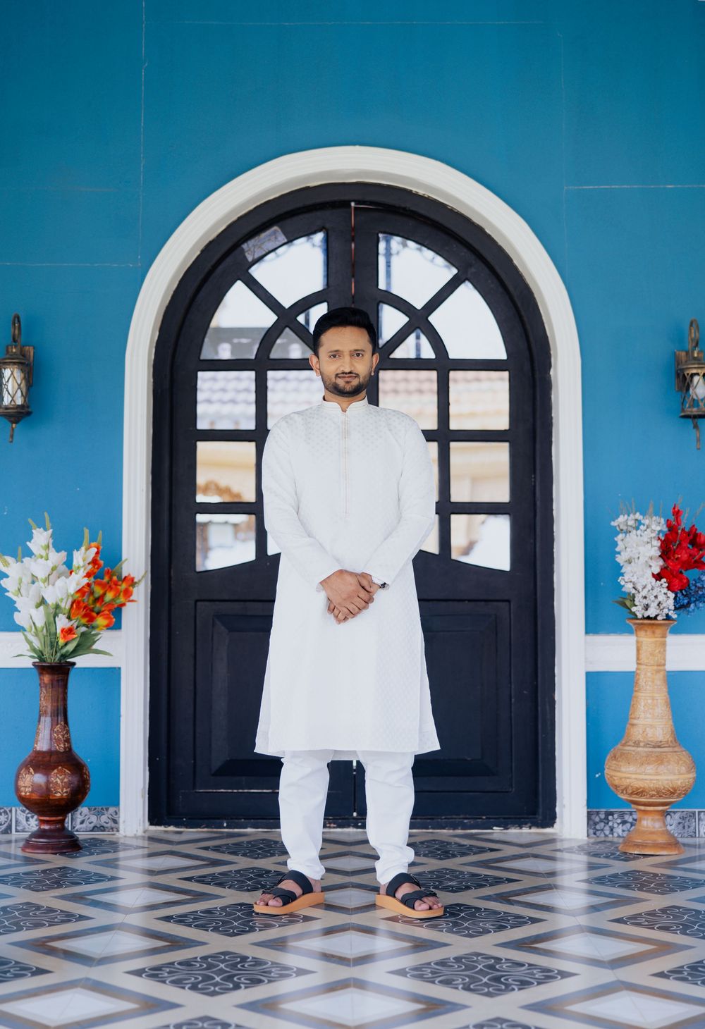 Man in white kurta stands before ornate black arched door on blue wall with floral arrangements.