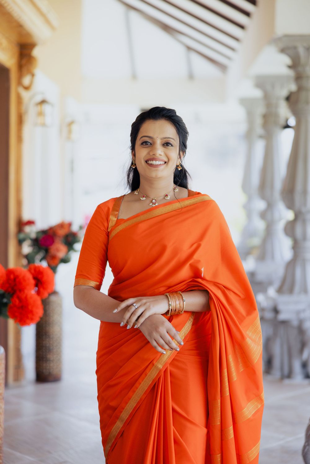 Smiling woman in vibrant orange saree with gold border and traditional jewelry poses in elegant corridor.