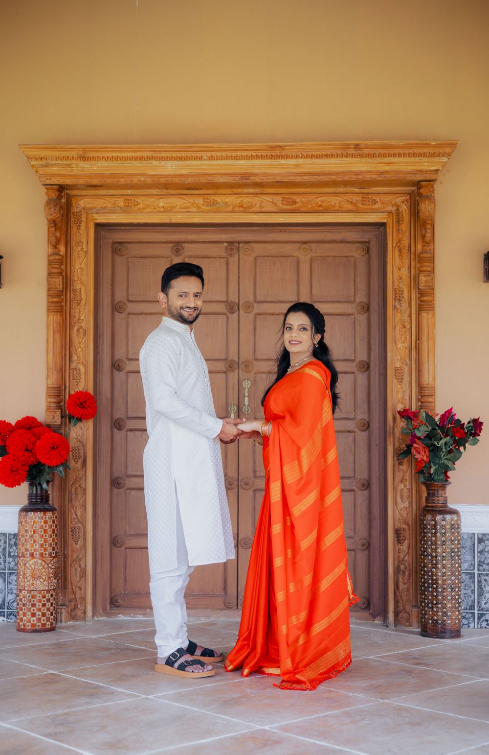 Couple in traditional Indian attire holding hands before ornate wooden doors with red floral arrangements.