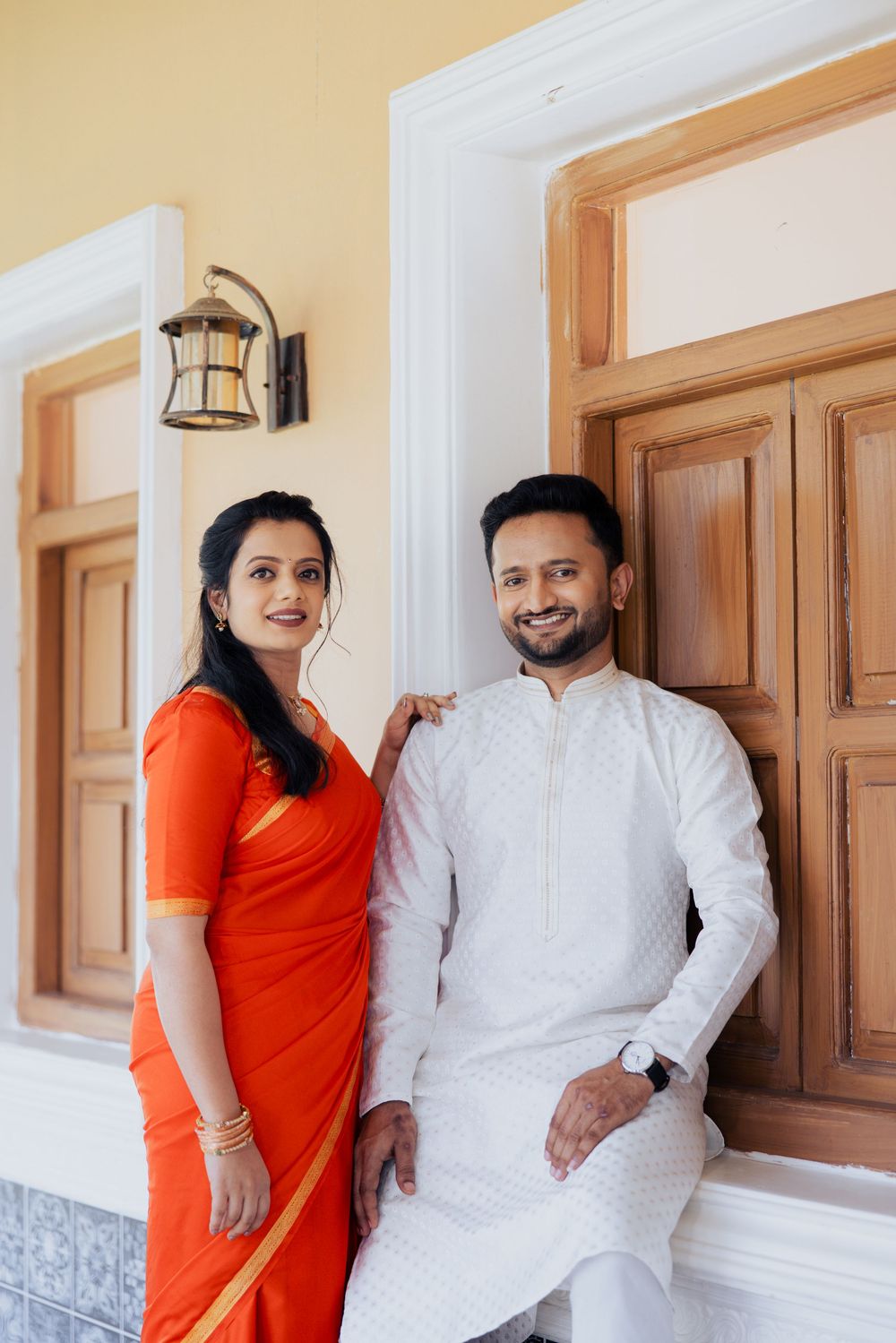 Indian couple in traditional attire, woman in orange saree and man in white kurta, posing near wooden doors.