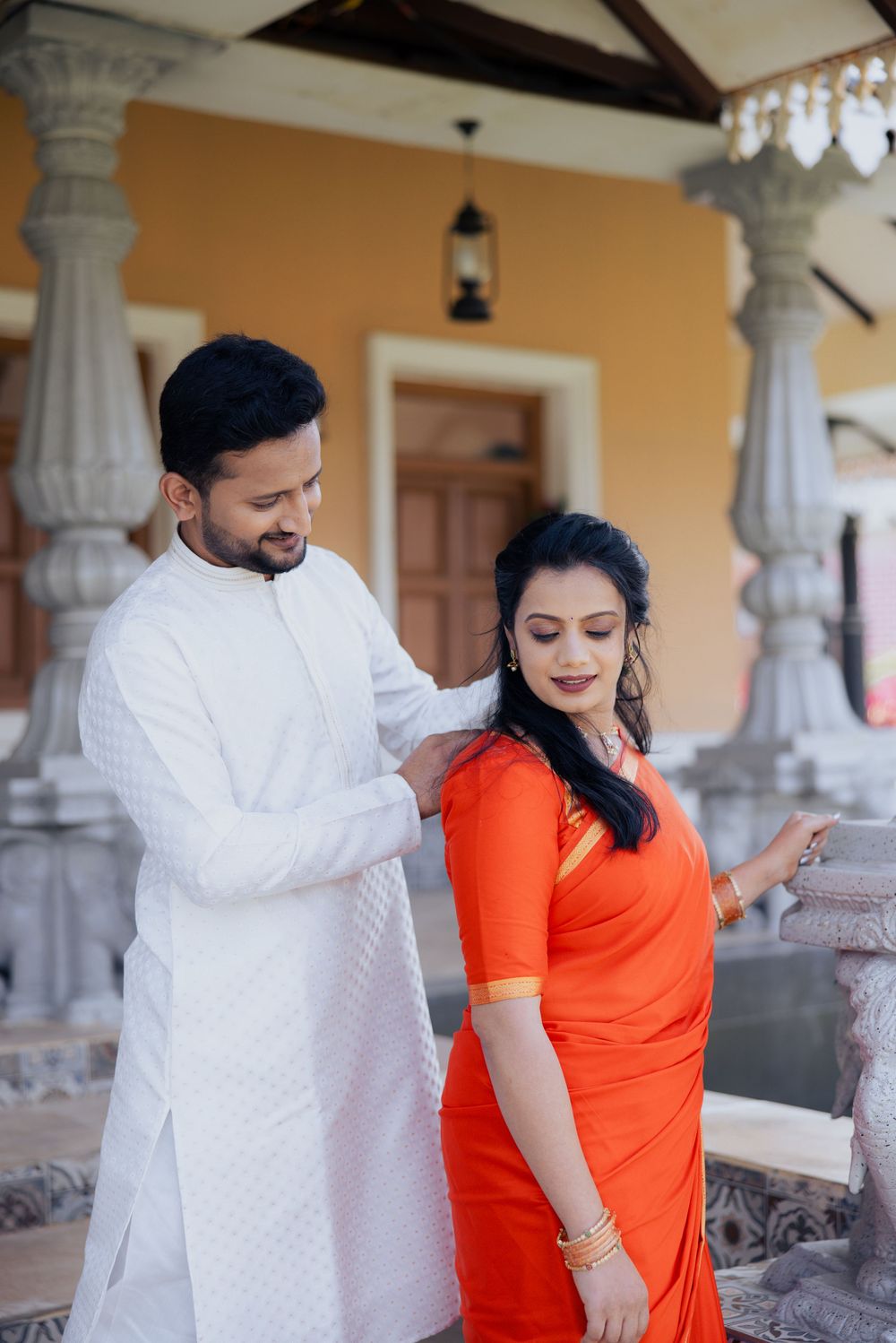 Indian couple in traditional attire, man in white kurta adjusting woman's orange saree near ornate stone columns.