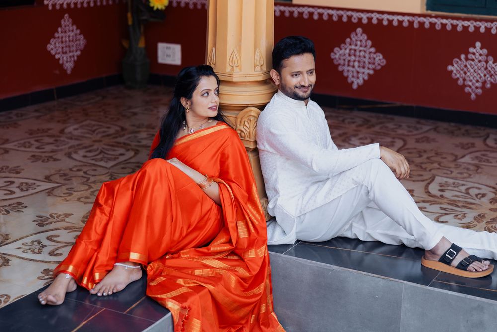 Indian couple in traditional attire, woman in orange saree and man in white kurta, sitting back-to-back in a temple.