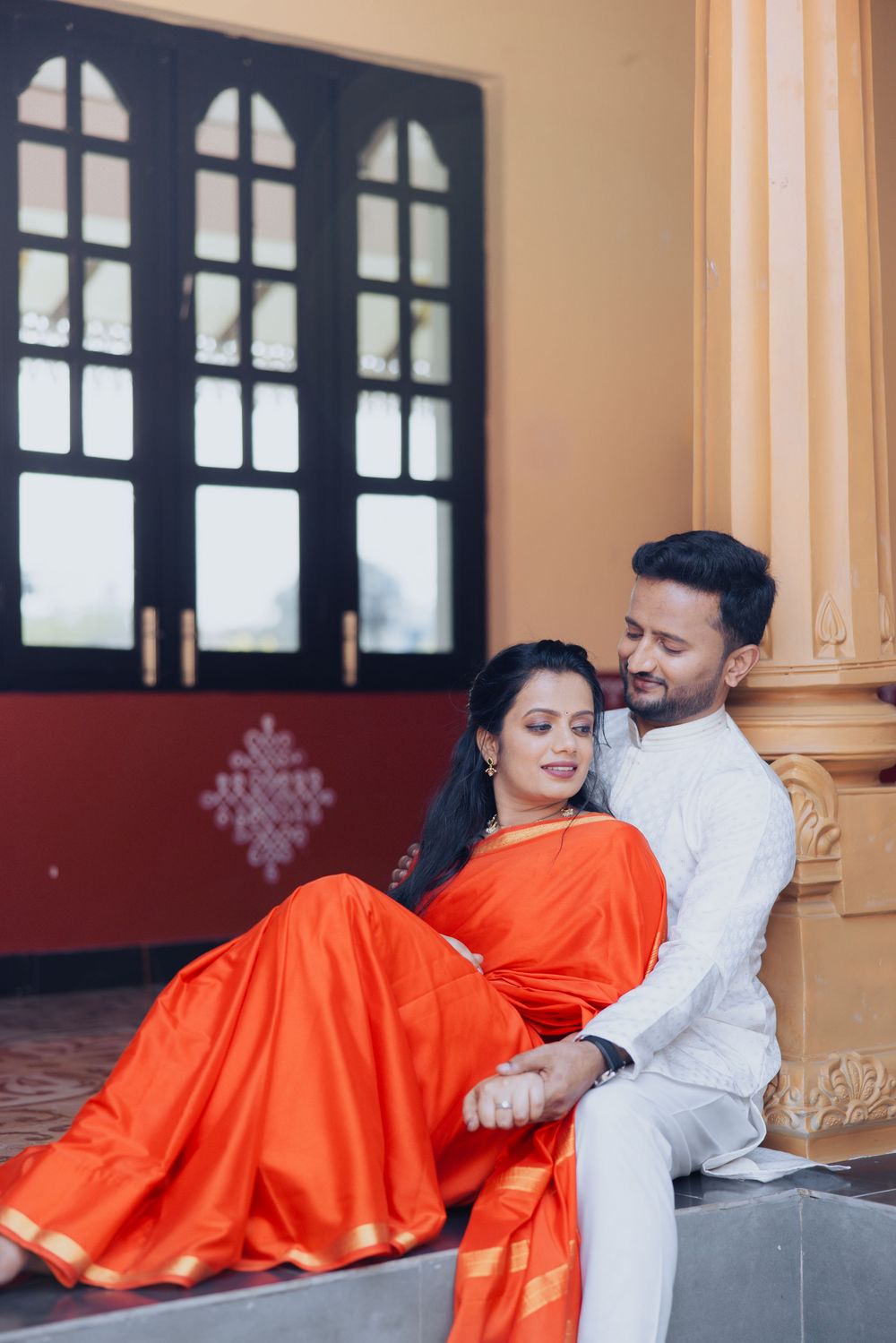 Indian couple in traditional attire, woman in orange saree and man in white kurta, sitting together near ornate pillars.