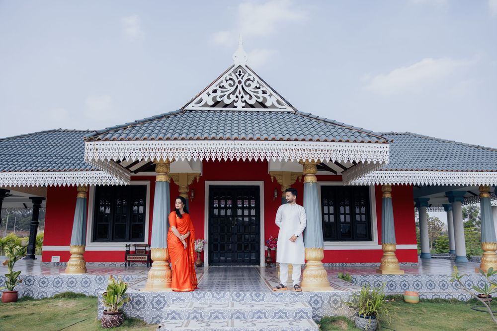 Couple in traditional Indian attire standing before a red and white heritage-style home with ornate roof and columns.