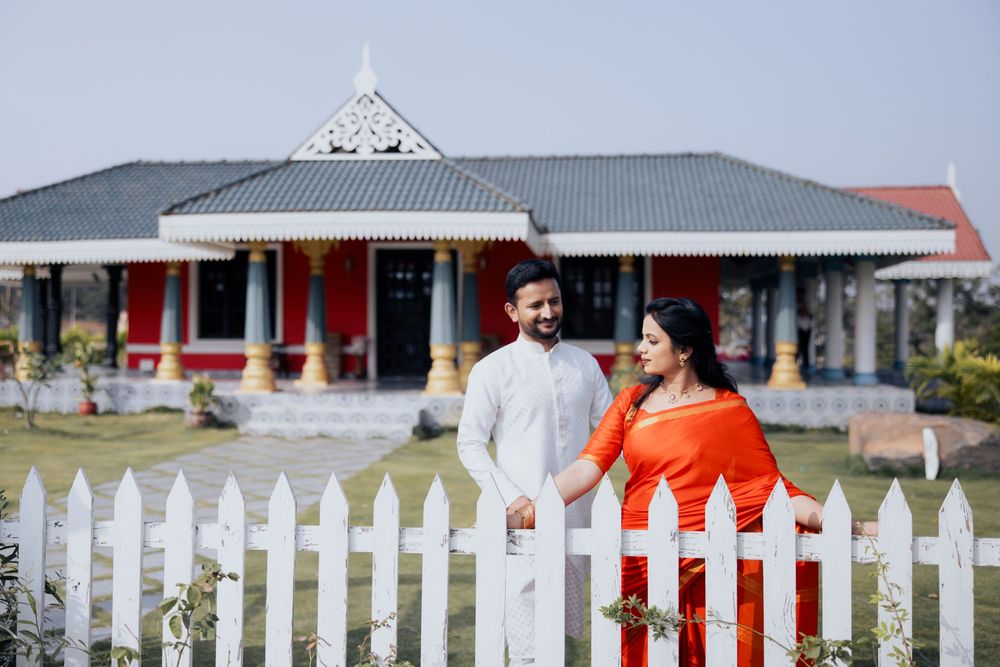 Couple in traditional attire holding hands by white picket fence with ornate building backdrop.