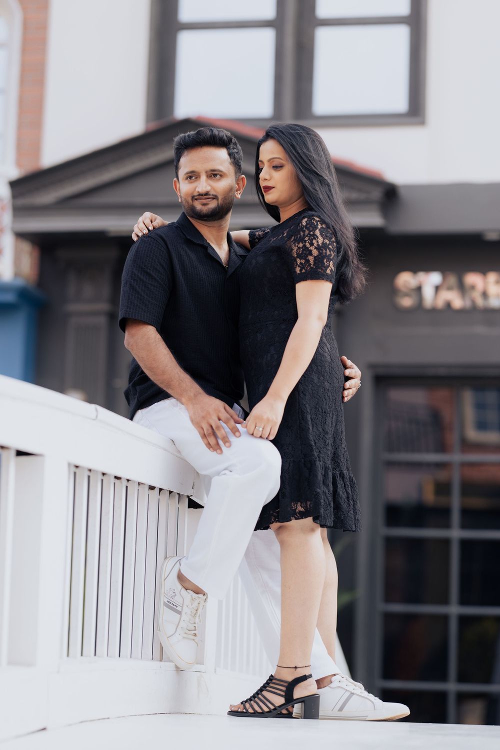 Couple in black outfits posing outdoors near a building, woman in lace dress and man in white pants.