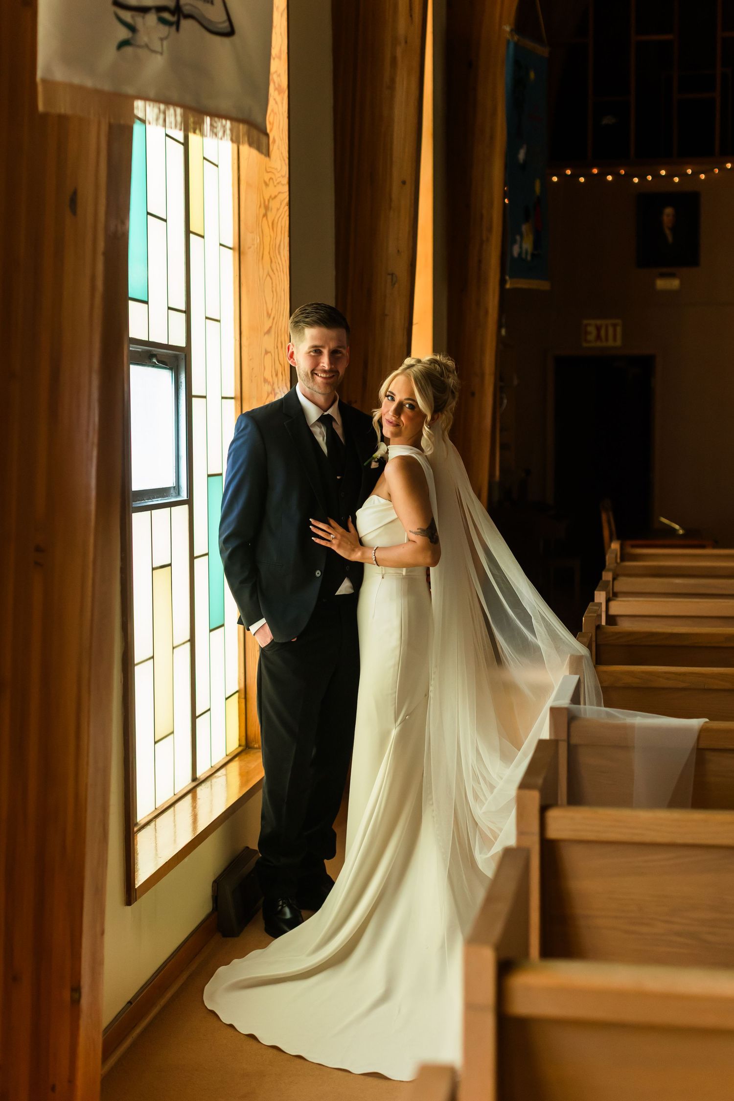 Bride in white gown and veil stands with groom in navy suit by stained glass windows inside a church.