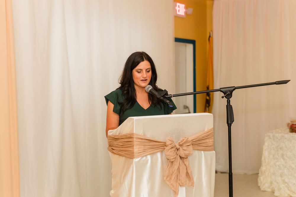 Woman in green dress speaking at a podium with microphone at an elegant event venue.