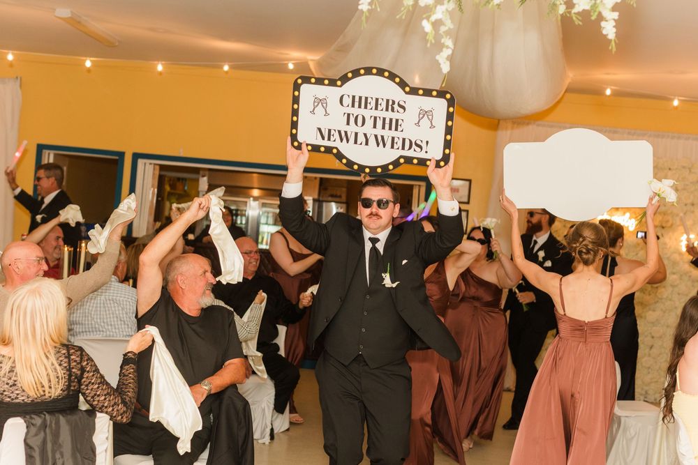 Wedding guests celebrate newlyweds, man holds 'Cheers to the Newlyweds' sign at lively indoor reception.