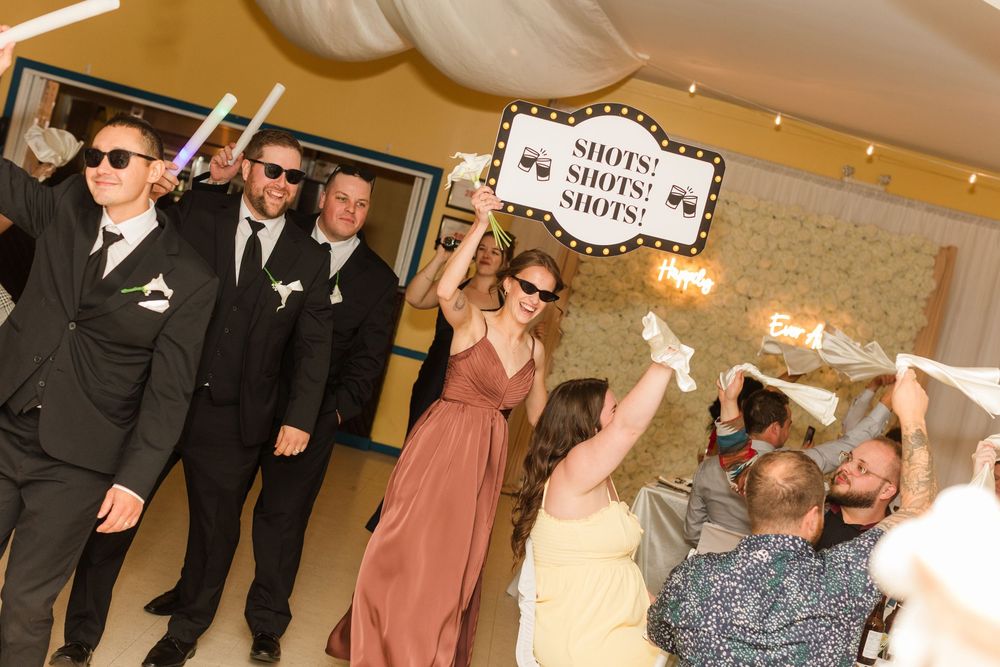 Wedding party celebrating indoors, groomsmen in suits and bridesmaid holding a Shots sign during lively reception festivities.