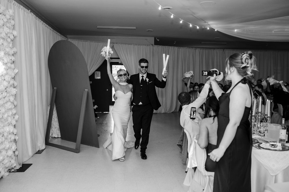 Bride and groom make grand entrance at wedding reception holding light wands while guests celebrate and photographer captures moment.