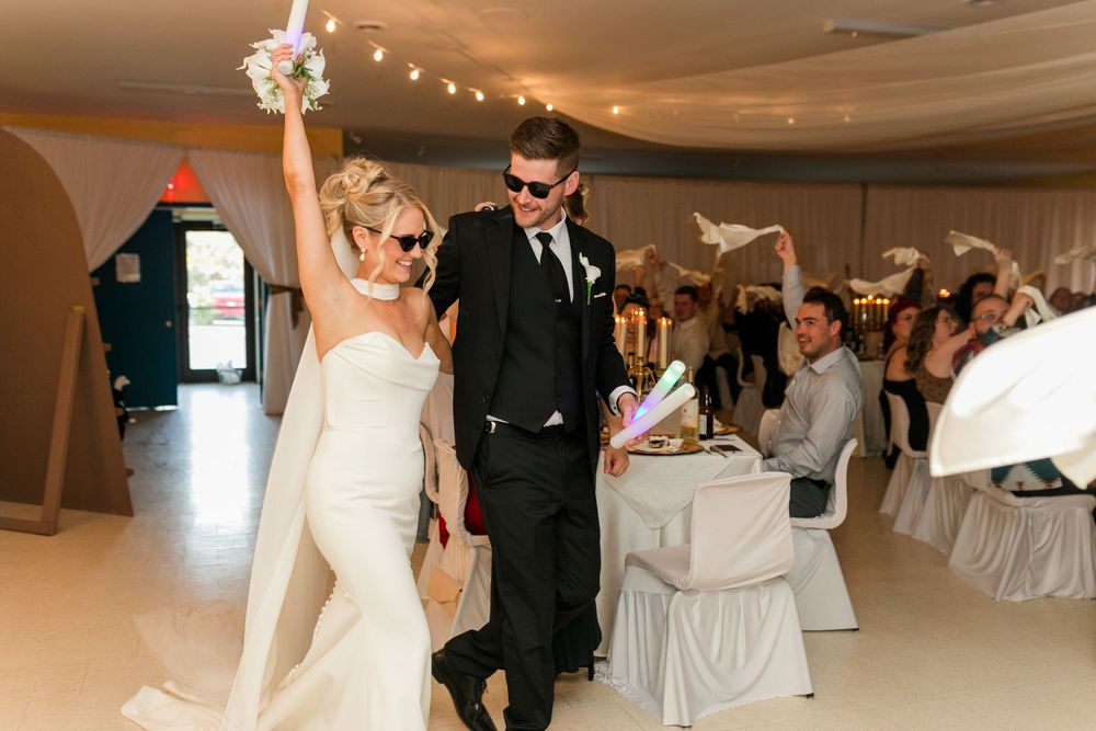 Bride and groom make grand entrance at wedding reception, bride raising bouquet, guests waving napkins in celebration.
