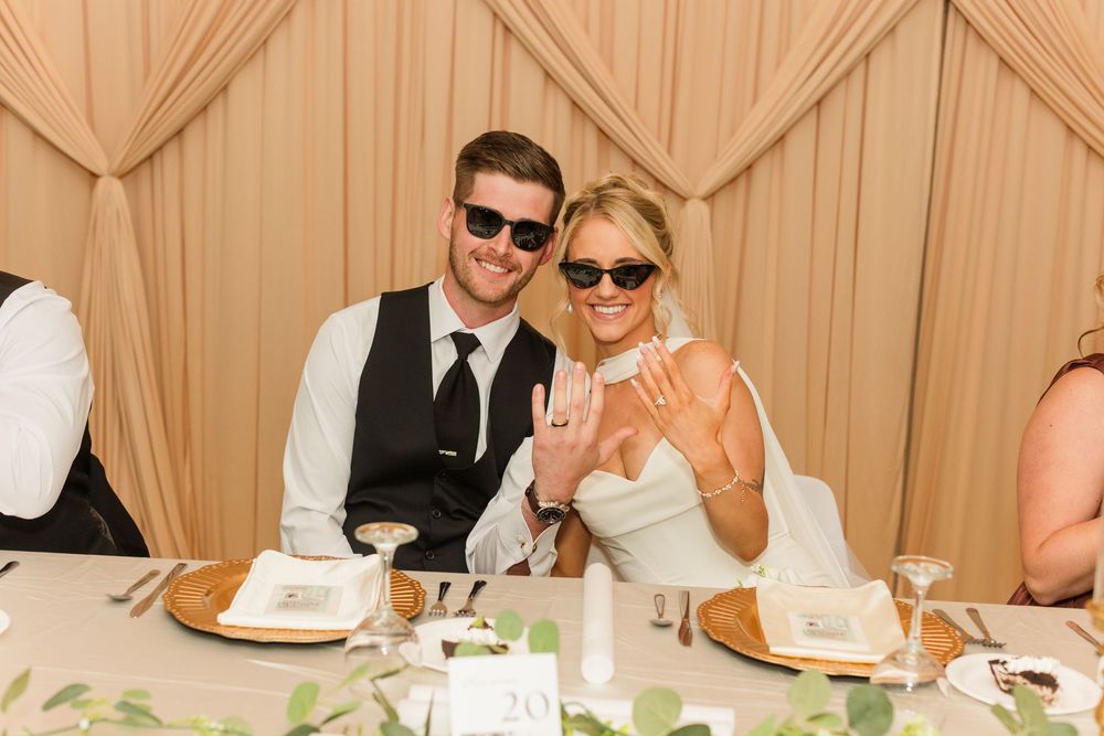 Newlywed couple in sunglasses showing wedding rings at reception table.