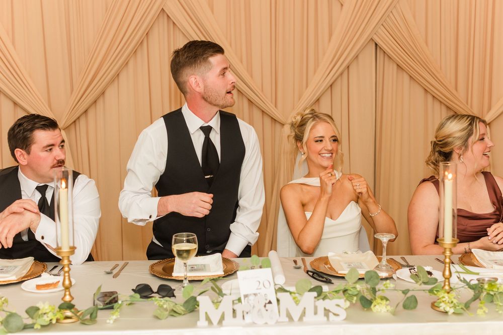 Bride and groom laughing at wedding reception head table with bridal party, decorated with greenery and Mr & Mrs sign.